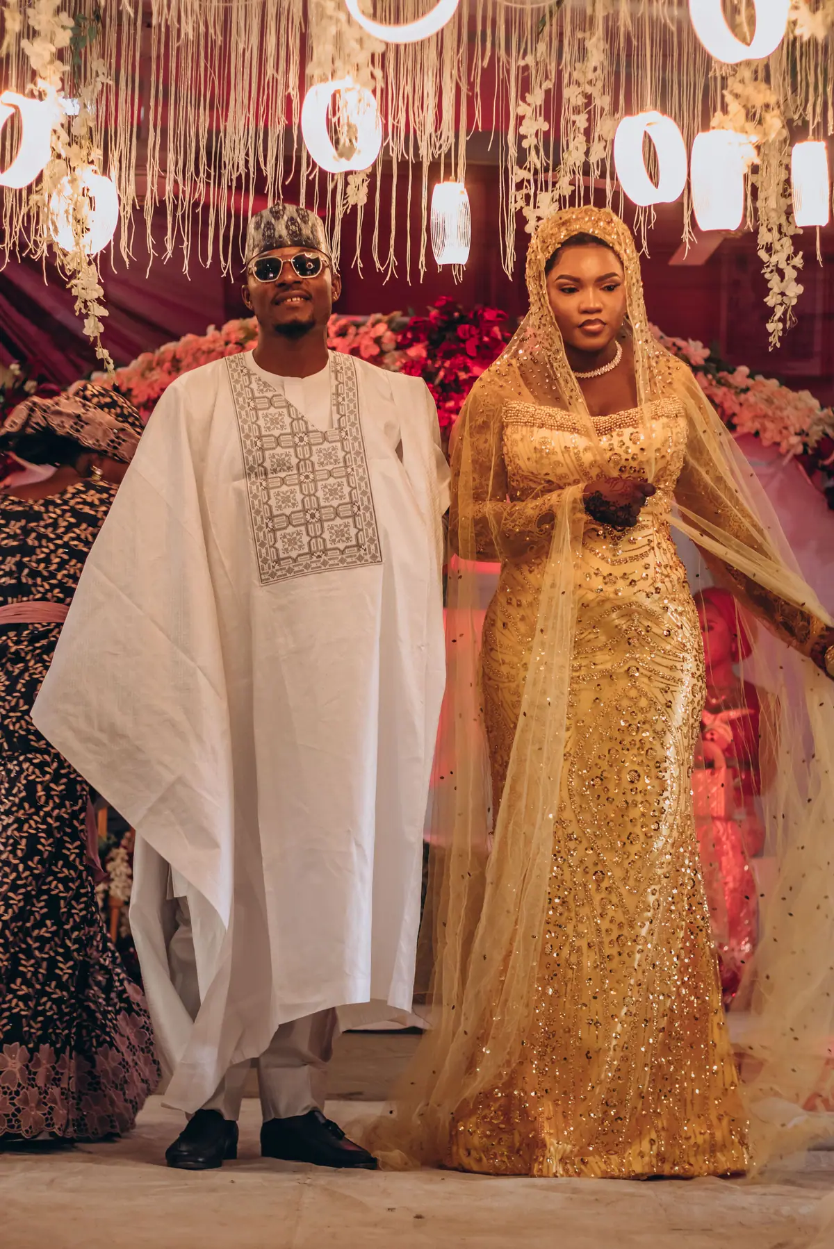A couple in white agbada and a gold traditional gown standing under floral event lighting.
