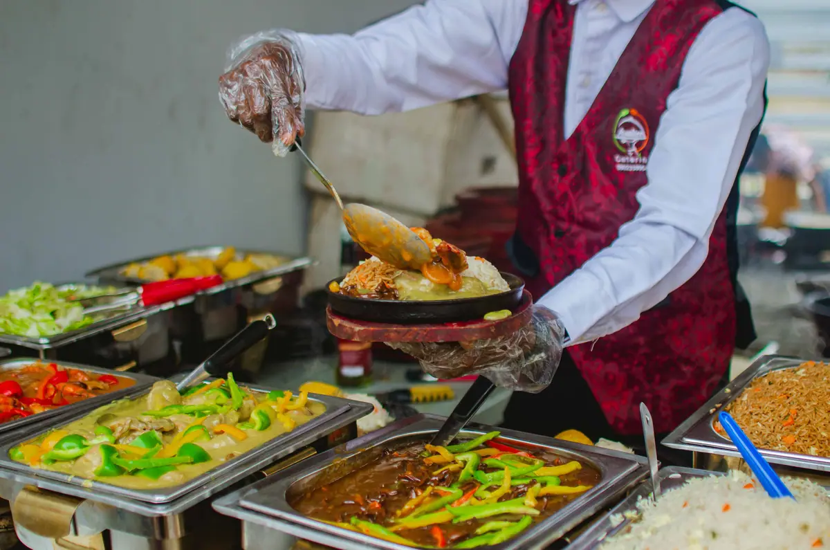 A caterer in gloves serving food from a buffet of chafing dishes.